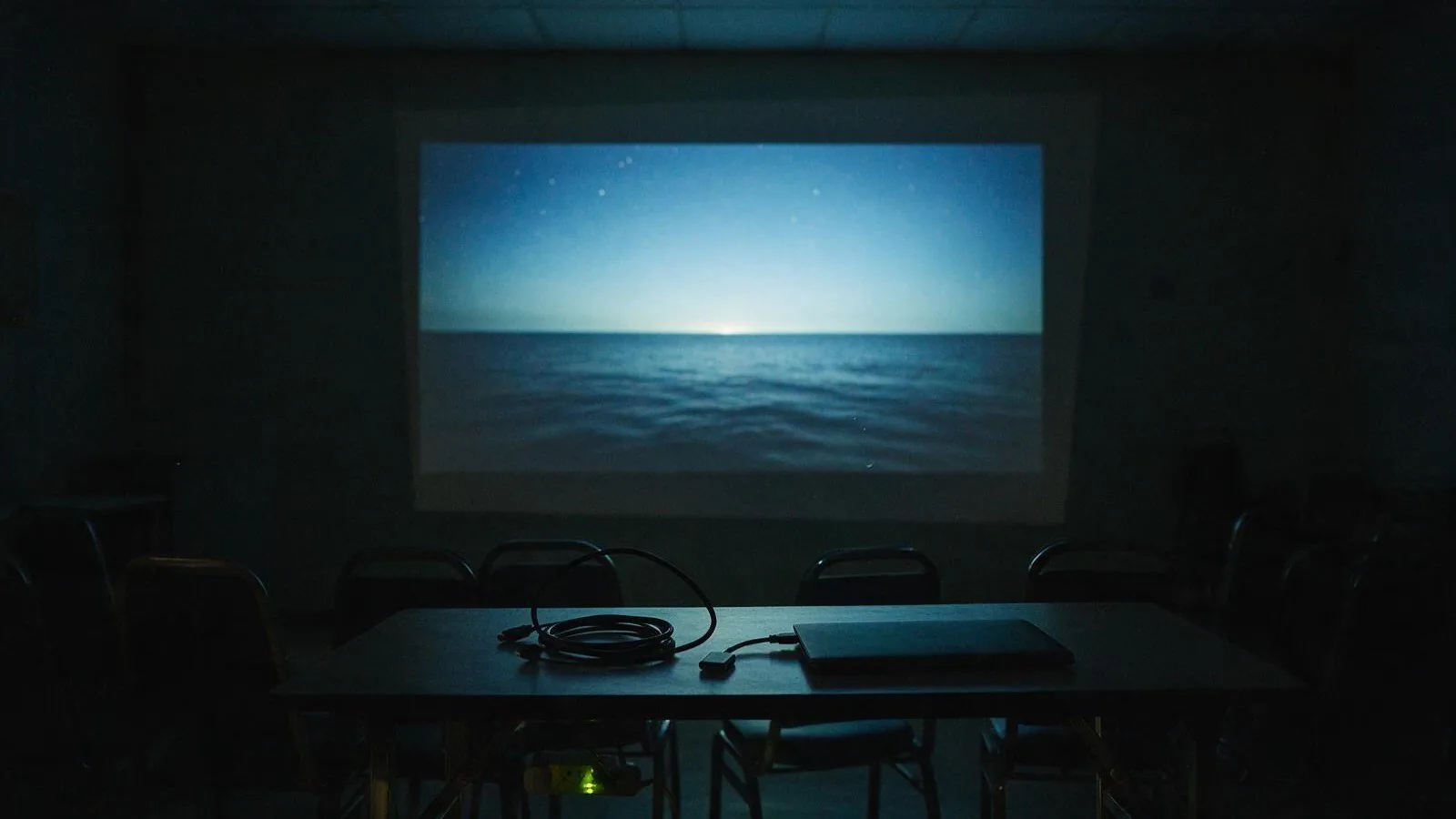 A dark training room at night with a projected seascape on the wall, a coiled HDMI cable, an adapter and a closed laptop on the table